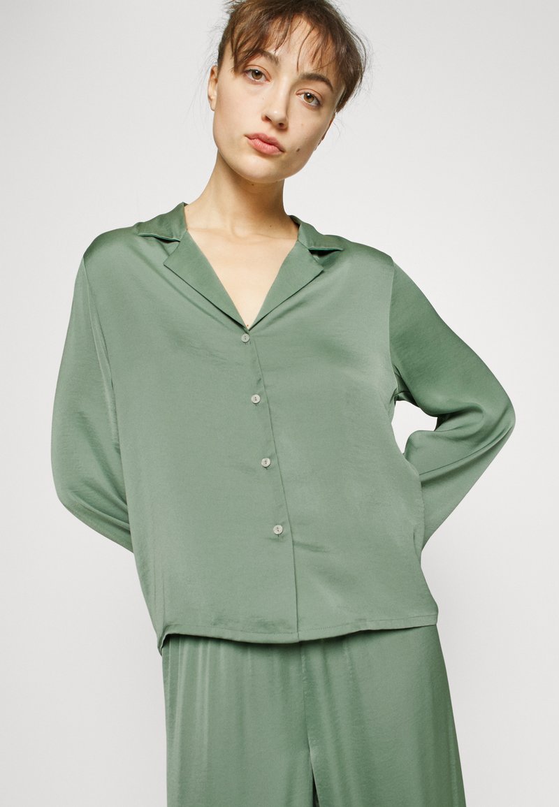Camicia verde in satin con colletto, maniche lunghe e vestibilità comoda. Presenta piccoli bottoni e una texture liscia e lucida.