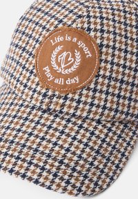 Barna, sötétkék és fehér houndstooth mintás sapka. Kerek barna folttal rendelkezik, amelyen fehér szöveg olvasható: "Az élet egy sport, játssz egész nap."