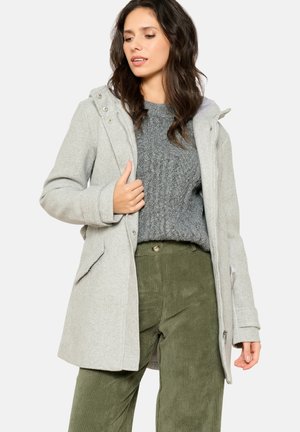 Manteau en mélange de laine gris clair avec une capuche, comportant une fermeture éclair et des boutons-pression, deux poches avant, superposé sur un pull en tricot gris et un pantalon en velours côtelé vert.