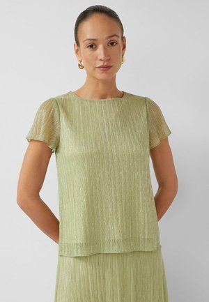 Vrouw met naar achteren gekamd haar, die gouden oorringen draagt en een doorschijnende lichtgroene geplooide top met korte mouwen en een bijpassende rok.