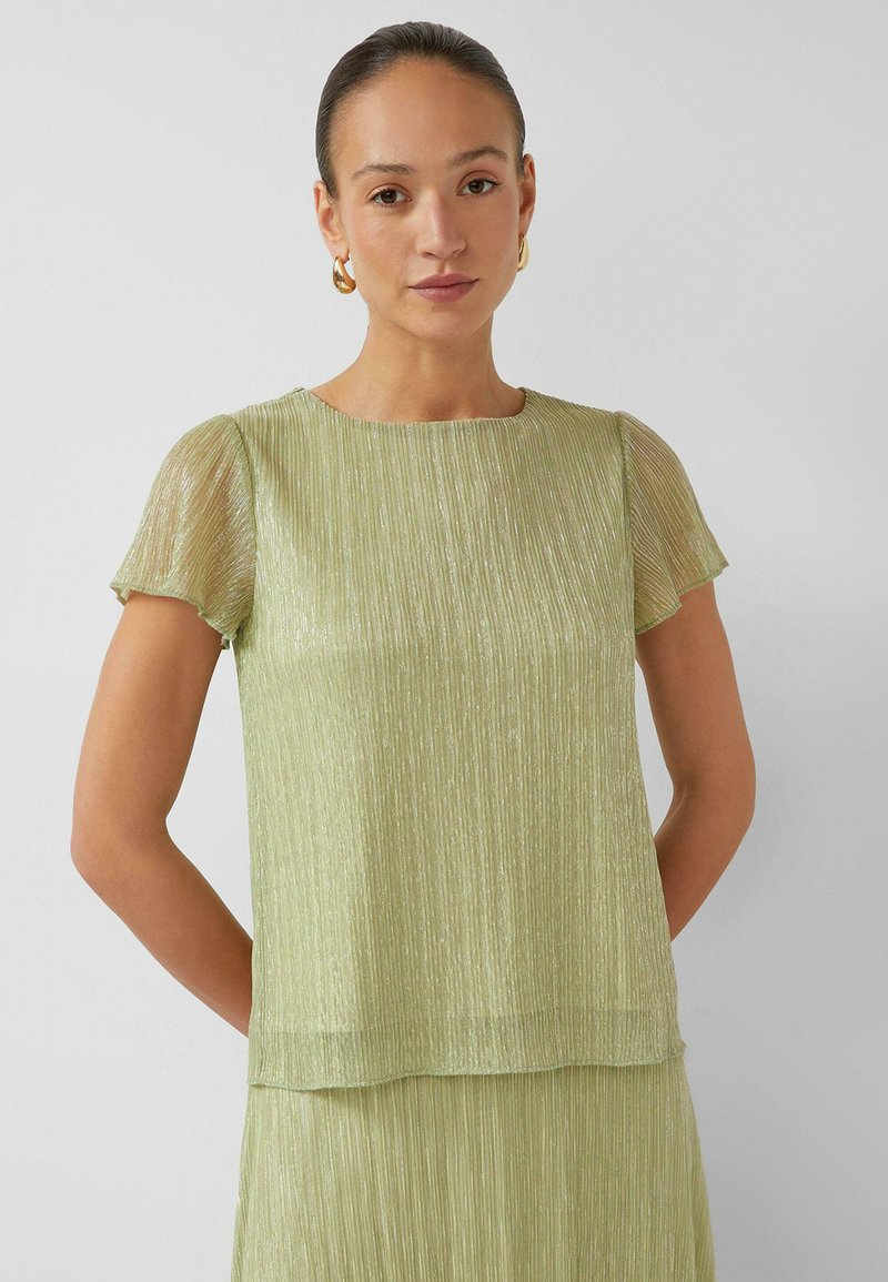 Vrouw met naar achteren gekamd haar, die gouden oorringen draagt en een doorschijnende lichtgroene geplooide top met korte mouwen en een bijpassende rok.