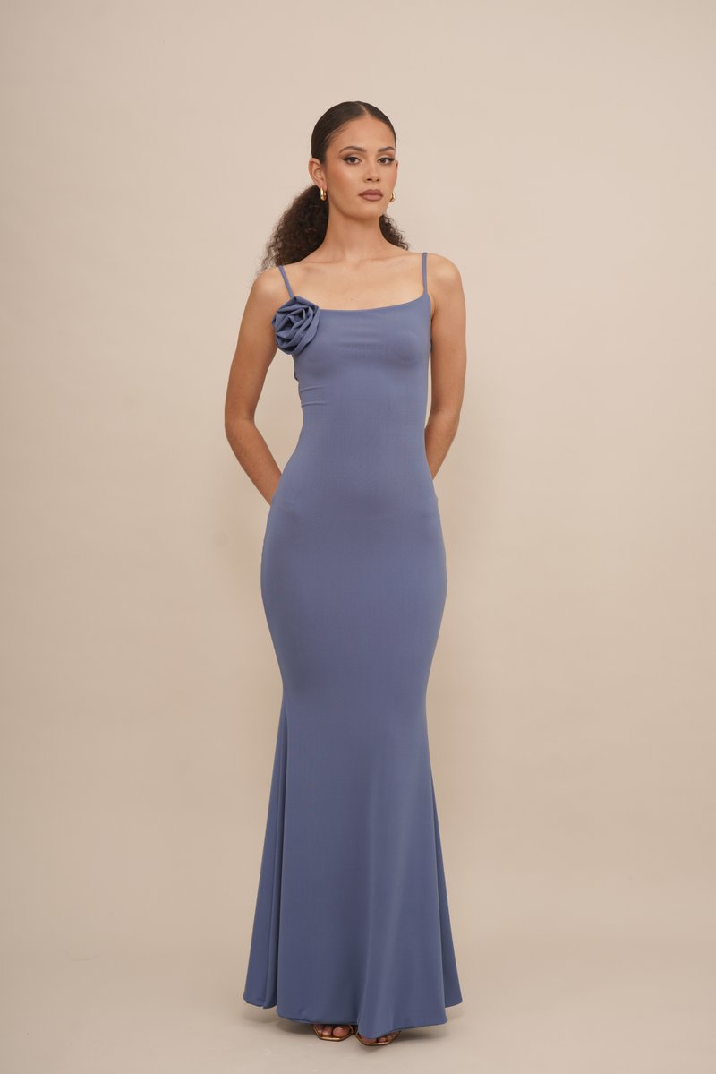 Vestido longo ajustado em material azul claro com um design de um ombro, apresentando um detalhe em rosa de tecido. Textura suave, estilo minimalista.