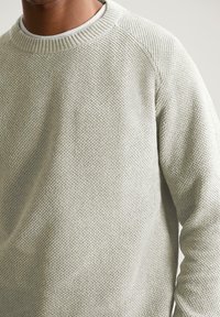 Svijetlosiva pletiva majica s teksturiranim uzorkom, okruglim izrezom i rebrastim rukavima. Ima ležeran kroj i raglan rukave.
