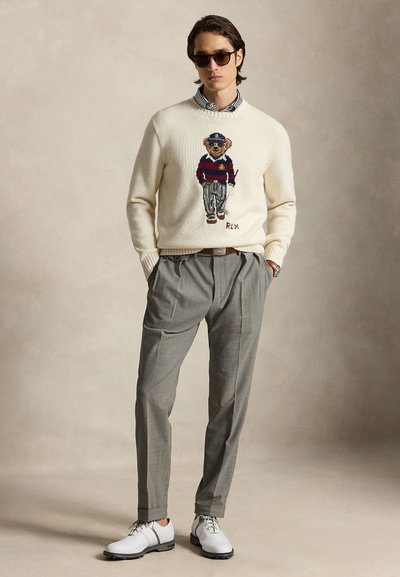Homme portant des lunettes de soleil, pull crème avec un motif d'ours en peluche, pantalon plissé gris, chaussures blanches, debout les mains dans les poches sur fond uni.