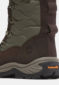 Dunkelbraune Wildleder- und olivgrüne Textilstiefel mit strukturiertem Wellenmuster, Metallösen und brauner Gummisohle mit Timberland-Logo.