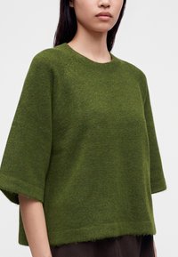 Femme portant un pull en laine vert oversize avec des manches jusqu'au coude et un col rond, sur un fond clair uni.
