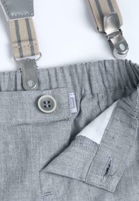 Un pantalon en denim gris présente une fermeture à bouton, une poche intérieure et des bretelles rayées beige et gris attachées avec des clips en métal.