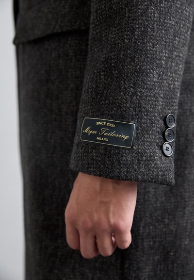 Cămașă de lână gri închis cu un model texturat, având un etichetă "Mgm Tailoring Milano" pe manșetă și trei nasturi negri.