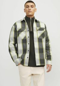Camicia in flanella a quadri grigia, bianca e verde lime, con bottoni frontali, tasche sul petto e un maglione verde scuro a strati sotto.