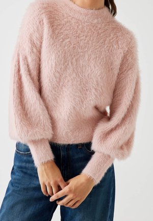 Femme portant un pull en maille pelucheuse rose clair avec des manches ballon et des poignets ajustés, assorti à un jean en denim bleu.