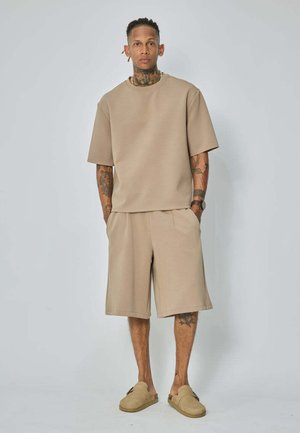 Homme avec des tatouages portant une chemise beige oversize à manches courtes, un short long assorti et des chaussures beiges sans lacets, debout devant un fond uni.