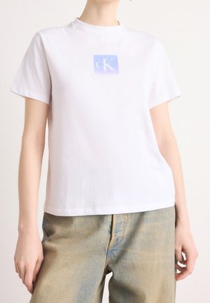 Persoon draagt een wit T-shirt met korte mouwen met een blauw vierkant logo en beige hoge taille jeans, staand tegen een effen lichte achtergrond.