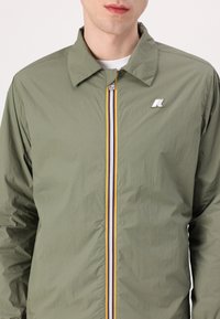 Olive green lightweight jacket with a front zipper featuring a multicolored stripe. Minimalist design with a logo on the left chest.