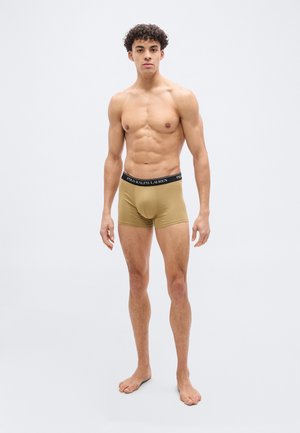 Junger muskulöser Mann steht barfuß und trägt beige Boxer-Shorts mit schwarzem Bund, auf dem "Polo Ralph Lauren" steht, vor einem einfarbigen hellen Hintergrund.