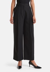 Černé kalhoty s širokými nohavicemi, sklady na přední straně, vysokým pasem a hladkým povrchem. Oblečené s lesklými černými lodičkami pro formální vzhled.