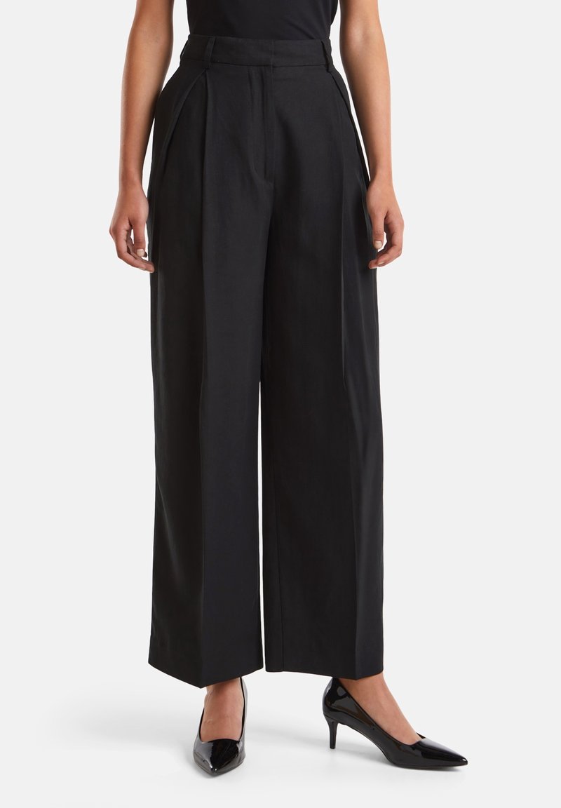 Černé kalhoty s širokými nohavicemi, sklady na přední straně, vysokým pasem a hladkým povrchem. Oblečené s lesklými černými lodičkami pro formální vzhled.