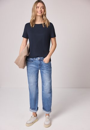 Mujer con camiseta azul marino, vaqueros azul claro con puños y zapatillas blancas, sosteniendo una chaqueta beige, de pie frente a un fondo claro y liso.