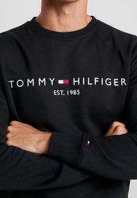 Sudadera negra con el logo bordado de "TOMMY HILFIGER" y el texto "EST. 1985". Los puños presentan un pequeño acento en rojo, blanco y azul.