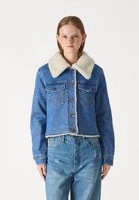 Przycięta niebieska kurtka dżinsowa z białym kołnierzem z owczej wełny, zapinana na guziki, z dwiema kieszeniami na piersi z pionowymi przeszyciami.