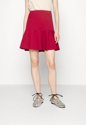 Femme portant une mini-jupe évasée rouge, un top beige et des chaussures plates texturées noir et blanc avec des liens à la cheville.