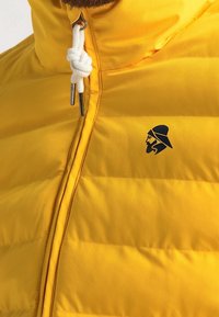 Gul vadderad jacka med hög krage, utrustad med en vit dragsko och metallkedja. Inkluderar en liten svart logotyp på bröstet.