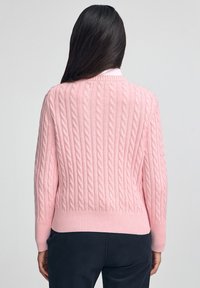 Kvinna med långt mörkt hår som bär en rosa kabelstickad tröja och mörka byxor, sedd bakifrån mot en enfärgad bakgrund.