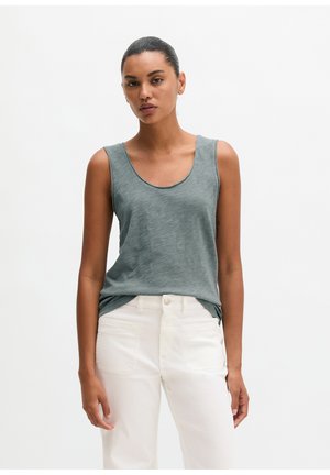 Frau mit zurückgebundenen Haaren, die ein ärmelloses graues Oberteil und weiße Hosen trägt und vor einem einfarbig weißen Hintergrund steht.