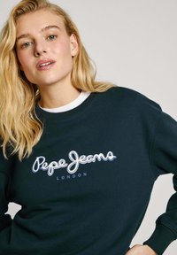 Jūras zilas krūšturiņu džemperis, kas izgatavots no kokvilnas maisījuma. Iezīmēts ar baltu "Pepe Jeans London" logotipu skripta fontā, ar ribotām piedurknēm un apkakli.