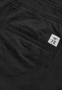 Črne tkanine hlače z zadnjim žepom, z belim označevalnikom "M" in "73". Gladka tekstura in čisti šivi so vidni.