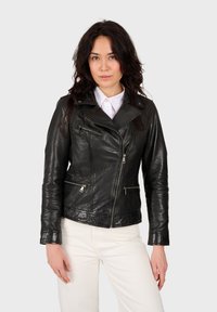 Veste de motard en cuir noir avec fermeture éclair asymétrique, coupe slim et deux poches zippées à l'avant, associée à une chemise blanche et un pantalon crème.