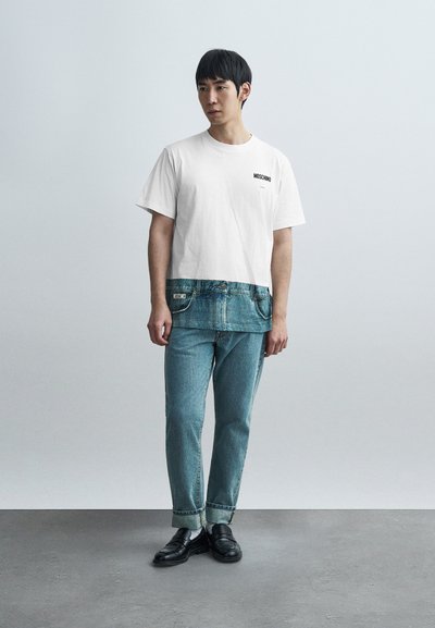 Λευκό κοντό μπλουζάκι με το λογότυπο "MOSCHINO", μπλε τζιν λεπτομέρειες στο τελείωμα. Ανοιχτό μπλε τζιν παντελόνια με γυρισμένα μανσέτα, μαύρα μοκασίνια.