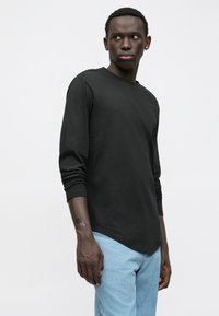 Mladý muž s tmavou pletí, oblečený v černé košili s dlouhým rukávem a světle modrých džínách, stojící na čistém bílém pozadí a dívající se bokem.