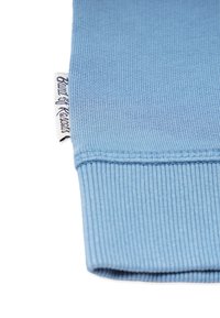 Helles blaues geripptes Strickbündchen mit einem weißen Stoffetikett, auf dem teilweise sichtbarer schwarzer Schrifttext steht.