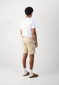 Un short cargo beige avec plusieurs poches latérales et une taille. Associé à un t-shirt blanc simple et des sabots marron clair avec des chaussettes blanches.
