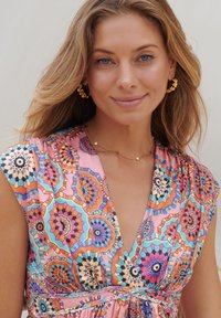 Colorful, patterned fabric dress with geometric motifs in pink, orange, and blue. Features a V-neckline and gathered waist. Gold hoop earrings.