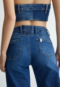 Bakvy av person som bär högmidjade blå denimjeans med insydd midja och matchande kort denimtopp.