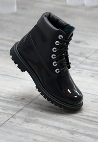 Stivaletto nero lucido con colletto imbottito, parte anteriore con lacci, accessori in argento e suola ruvida e texture. Presenta un design moderno e raffinato.