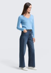 Helles blaues V-Ausschnitt-Sweatshirt mit langen Ärmeln, kombiniert mit hochgeschnittenen, dunkelblauen weitem Jeans. Weiße Sneaker runden den Look ab.