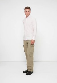 Φωτεινό ροζ πουκάμισο με κουμπιά και κλασικό γιακά, συνδυασμένο με μπεζ παντελόνι cargo με πολλαπλές τσέπες. Μαύρα παπούτσια ολοκληρώνουν το σύνολο.