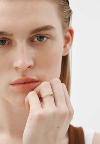 Anillo de oro con tres pequeñas piedras verdes, llevado en un dedo. La modelo tiene la piel clara y el cabello claro, mostrando el anillo contra un fondo neutro.