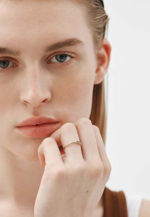 Anillo de oro con tres pequeñas piedras verdes, llevado en un dedo. La modelo tiene la piel clara y el cabello claro, mostrando el anillo contra un fondo neutro.