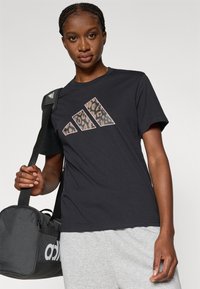 Črna bombažna majica s tiskanjem tigrovega vzorca in trakom s tremi črtami na sprednji strani. Model drži sivo športno torbo z belim logotipom.