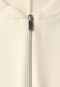 Sudadera cremera con cremallera plateada. La tela es suave y acanalada, cuenta con capucha y un diseño ajustado.