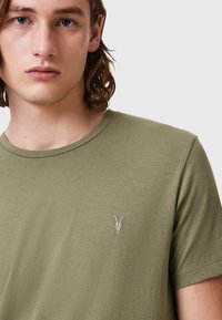 T-shirt in cotone verde con colletto a girocollo, caratterizzato da un piccolo emblema a skull ricamato sul lato sinistro del petto. Texture liscia, maniche corte.