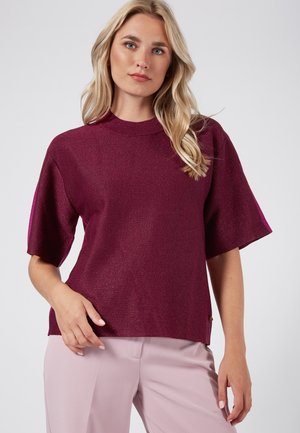 Burgundy knit top with short sleeves, ribbed neckline, and subtle shimmer. Paired with light pink trousers, showcasing a relaxed fit.