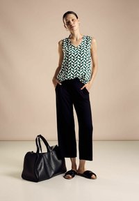 Top senza maniche con motivo geometrico verde, pantaloni larghi neri, borsa a tracolla in pelle nera e sandali neri su uno sfondo chiaro.