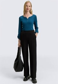 Turkizna majica Henley z dolgimi rokavi in tremi gumbi, kombinirana z visokim pasom in širokimi črnimi hlačami. Črna torbica čez ramo in masivni čevlji.