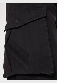 Black cargo pants made of durable fabric, featuring a side pocket with flap and fine diagonal ribbing texture.