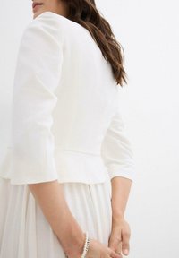 Giacca bianca strutturata con maniche a tre quarti, abbinata a un vestito bianco fluente. Tessuto morbido, dettaglio peplum sottile in vita.