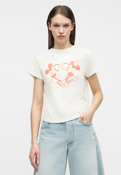 Νεαρή γυναίκα με ίσια μαλλιά που φοράει ένα λευκό κοντό T-shirt με πορτοκαλί Mickey και Minnie Mouse να κρατιούνται χέρι-χέρι, συνδυασμένο με ανοιχτό μπλε τζιν.